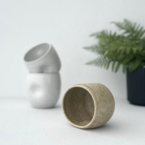 May include: A speckled beige ceramic mug with a rounded shape. The mug is sitting on a white surface. Two white ceramic mugs are stacked behind the beige mug.