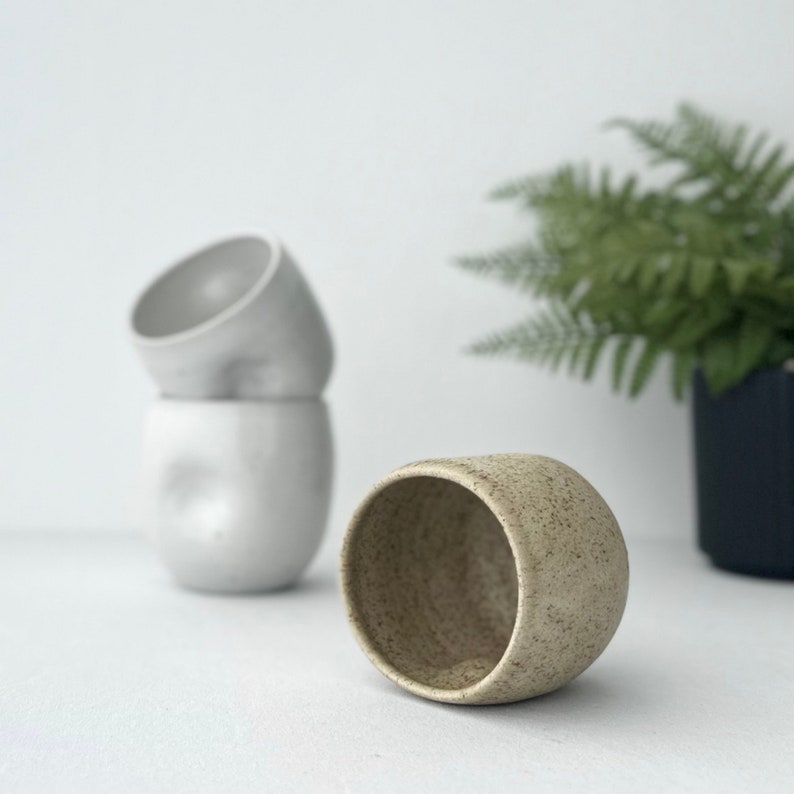 May include: A speckled beige ceramic mug with a rounded shape. The mug is sitting on a white surface. Two white ceramic mugs are stacked behind the beige mug.