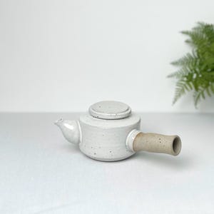 May include: A small, white ceramic teapot with a round lid and a side handle. The teapot has a spout and a speckled texture. The handle is a light brown colour. The teapot is designed for brewing tea.
