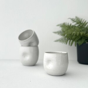 May include: Three white ceramic mugs with a textured, uneven surface. The mugs are stacked on top of each other, with the top mug tilted slightly. The mugs are sitting on a white surface.