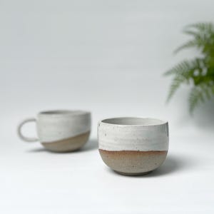May include: Two handmade ceramic mugs. One has a handle, the other is bowl-shaped. Both are white with a brown band at the base. The mugs are on a white surface.