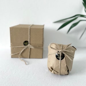 May include: Two brown paper wrapped packages tied with twine. One package is a square box and the other is a round container. Both packages have a black and gold sticker with text.