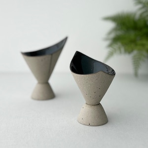 May include: Two small, ceramic, cone-shaped bowls with black interiors and speckled, light grey exteriors. The bowls are on a white surface.