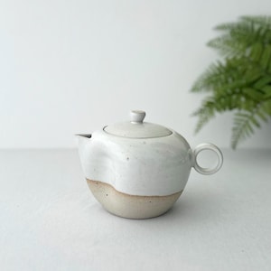 Op de afbeelding: Een witte theepot van keramiek met een gespikkeld glazuur en een bruine basis. De theepot heeft een rond lichaam en een kleine tuit.