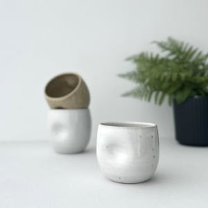 May include: Three ceramic mugs, two are brown and one is white. The white mug has a textured surface and is in focus. The brown mugs are out of focus and stacked on top of each other.