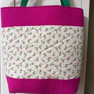 May include: A quilted tote bag with a fuchsia top and bottom, and green handles. The central panel is cream-coloured fabric with a delicate floral pattern in pink and green, stitched in a diamond pattern.