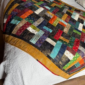 May include: A vibrant patchwork quilt featuring a black base and rectangular fabric pieces in red, orange, green, blue, and gold. The quilt has a gold border and is displayed on a white bedspread.