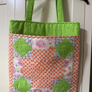 May include: A quilted tote bag with green polka dot handles and top border. The main body features a floral pattern with orange and white flowers, accented by green diamond shapes. The bag is designed for carrying items.