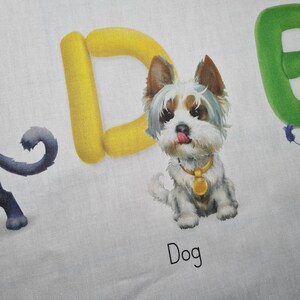May include: A white fabric with a colorful illustration of a dog and the word "Dog" in black text. The dog is a small, fluffy breed with a gold collar and a pink tongue sticking out. The letter "D" is yellow.
