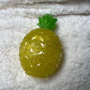 May include: A yellow pineapple-shaped ornament with a green crown. The pineapple has a textured, geometric surface. The object is displayed on a white, fluffy background.