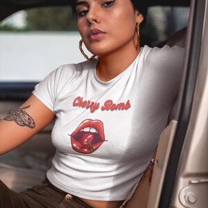May include: White crop top with the text "Cherry Bomb" in red script above a graphic of red lips with a cherry. The shirt is worn with brown pants. The person is sitting in a car.