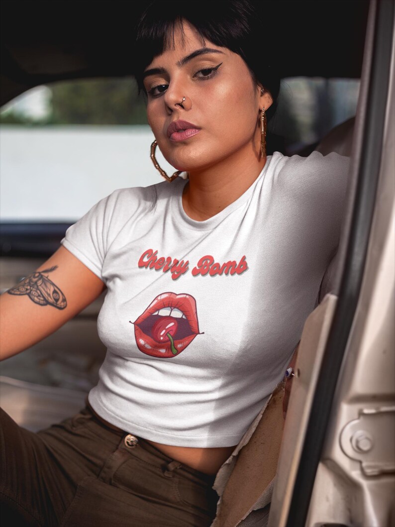 May include: White crop top with the text "Cherry Bomb" in red script above a graphic of red lips with a cherry. The shirt is worn with brown pants. The person is sitting in a car.
