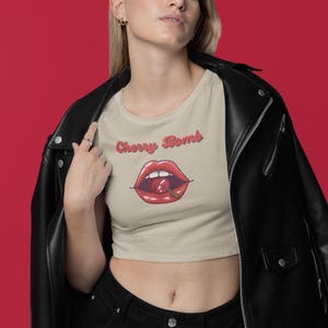 May include: Tan crop top with the words "Cherry Bomb" above a graphic of red lips with a cherry. The model is wearing a black leather jacket and black jeans. The background is red.