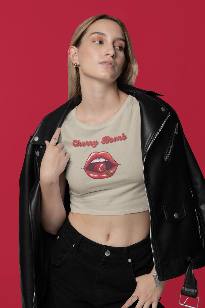 May include: Tan crop top with the words "Cherry Bomb" above a graphic of red lips with a cherry. The model is wearing a black leather jacket and black jeans. The background is red.