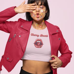 May include: A person wearing a red leather jacket and a light pink crop top with the text "Cherry Bomb" and a graphic of red lips with a cherry. The person has stickers on their face.
