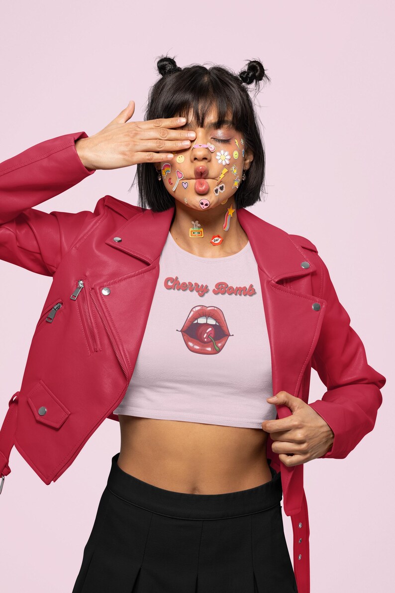 May include: A person wearing a red leather jacket and a light pink crop top with the text "Cherry Bomb" and a graphic of red lips with a cherry. The person has stickers on their face.