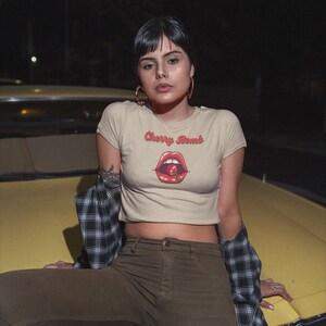 May include: A beige crop top with the text "Cherry Bomb" above a graphic of red lips and a cherry. The person is wearing brown pants and a black and white plaid shirt.