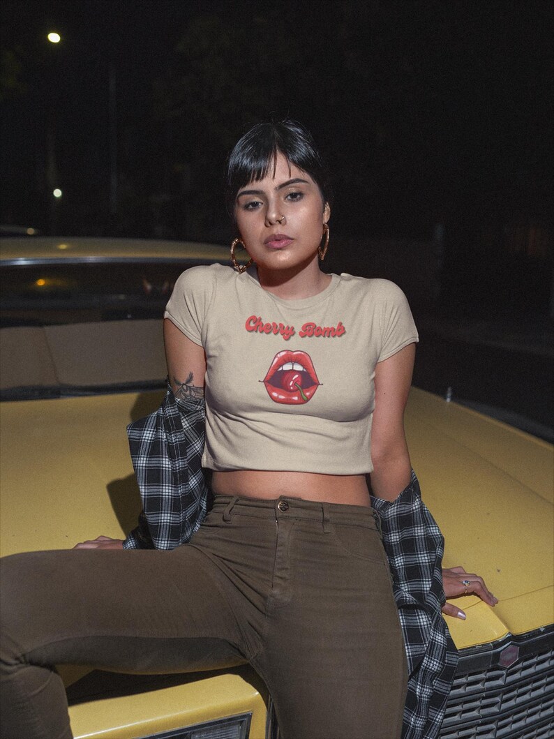 May include: A beige crop top with the text "Cherry Bomb" above a graphic of red lips and a cherry. The person is wearing brown pants and a black and white plaid shirt.
