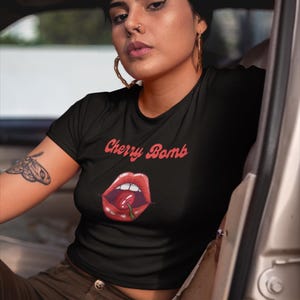 May include: Black t-shirt with the text "Cherry Bomb" in red script above a graphic of red lips with a cherry. The shirt is short-sleeved.