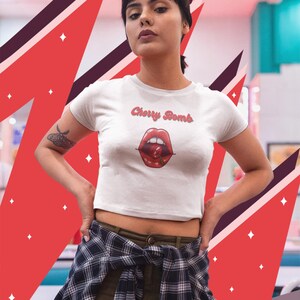 May include: White crop top with the text "Cherry Bomb" above a graphic of red lips and a cherry. The shirt is short-sleeved and has a round neckline. The background features red and maroon geometric shapes.