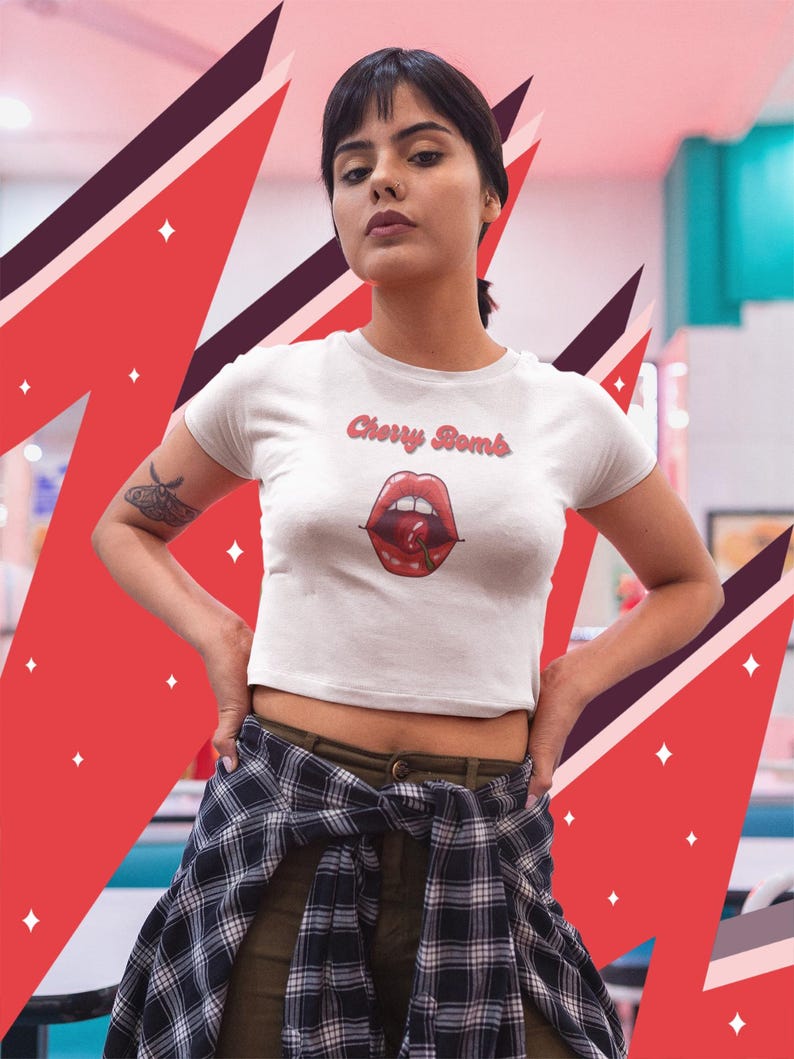 May include: White crop top with the text "Cherry Bomb" above a graphic of red lips and a cherry. The shirt is short-sleeved and has a round neckline. The background features red and maroon geometric shapes.