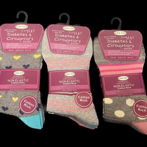 Op de afbeelding: Drie paar grijze, roze en witte sokken met een label met de tekst "For Her Diabetes & Circulatory Socks Non Elastic Cotton Rich".