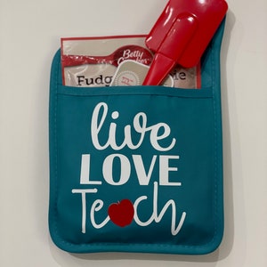 May include: Teal blue oven mitt with the text "live love teach" in white with a red apple in the middle of the word "teach". The oven mitt has a pocket with a red spatula and a Betty Crocker fudge mix packet inside.