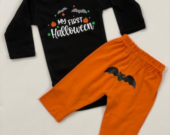 Może przedstawiać: Czarny body z długim rękawem i pomarańczowe spodnie dla dziecka. Body ma napis "MY FIRST Halloween" z nietoperzami i dyniami. Pomarańczowe spodnie mają grafikę nietoperza z tyłu.