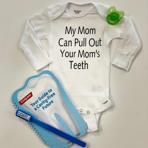May include: A white baby onesie with the text "My Mom Can Pull Out Your Mom's Teeth". A green pacifier and a blue toothbrush with a spiral bound book shaped like a tooth with the text "Colgate Your Guide to a Cavity-Free Future" are also in the image.