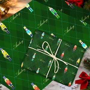 May include: Green wrapping paper with a repeating pattern of nutcracker soldiers and the name "Audrey" in a script font. A wrapped gift with the same design is tied with twine. The background is a marble surface with Christmas decorations.