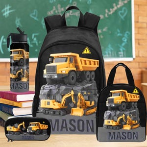 May include: A black backpack, lunch bag, pencil case, and water bottle set featuring yellow construction vehicles. The name "MASON" is printed on each item. The water bottle is next to a stack of books.