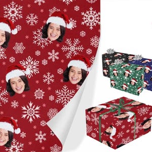 May include: Red wrapping paper with a white snowflake pattern and a repeated image of a person wearing a Santa hat. The paper is rolled up and the edges are visible. There are three wrapped boxes in the background, one green, one black, and one blue.