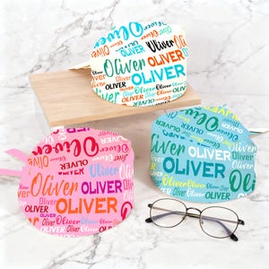 May include: Three colorful scrub caps with the name "Oliver" printed repeatedly in various fonts and colors. One cap is pink, another is blue, and the third is white. A pair of round-framed glasses rests in front of the caps.