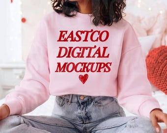 May include: A light pink sweatshirt with the text "EASTCO DIGITAL MOCKUPS" in red, along with a red heart graphic. The sweatshirt is paired with light blue denim jeans. The overall aesthetic is casual and comfortable.