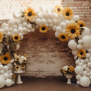 May include: A white balloon arch decorated with yellow sunflowers, greenery, and white flowers. The arch is in front of a brick wall. There is a white cake stand with a white cake on it in front of the arch.