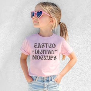Könnte beinhalten: Ein pinkfarbenes T-Shirt mit dem Text "EASTCO DIGITAL MOCKUPS" in einer Retro-Schriftart. Das Shirt wird von einem Kind getragen, das eine rosafarbene Herz-Sonnenbrille und eine blaue Jeans trägt.