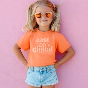 May include: A young person wearing a bright orange t-shirt with the text "east co digital mockups" printed on the front. The person is wearing blue denim shorts and has their hands on their hips. They are standing in front of a pink wall and wearing sunglasses.