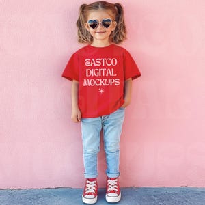 Può includere: Una persona giovane indossa una maglietta rossa con il testo "EASTCO DIGITAL MOCKUPS" in bianco. Indossa jeans blu e scarpe da ginnastica rosse. La persona è in piedi davanti a un muro rosa e indossa occhiali da sole a forma di cuore.