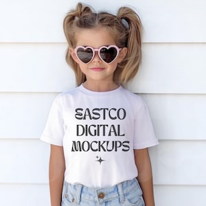 May include: A young person wearing pink heart-shaped sunglasses and a white t-shirt with the text "EASTCO DIGITAL MOCKUPS" in black. The person is also wearing blue denim shorts.