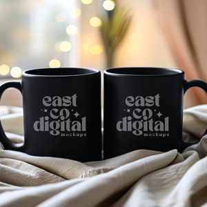 May include: Two black ceramic mugs with white text that reads "east co digital mockups" on a white fabric background.