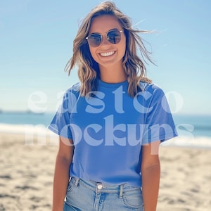 May include: A woman wearing a light blue t-shirt with a relaxed fit and a casual look. She is standing on a sandy beach with a blue sky and ocean in the background.