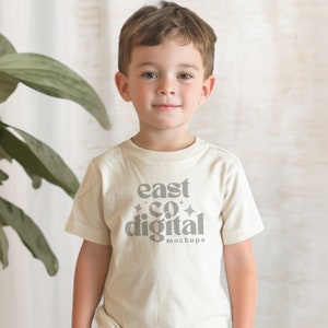 以下が含まれることがあります： 白いTシャツを着た小さな子供が、前面に「east co digital mockups」という文字が印刷されています。