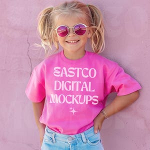 Może przedstawiać: Młoda osoba w jaskraworóżowej koszulce z białym napisem "EASTCO DIGITAL MOCKUPS". Ma również na sobie jasnoniebieskie dżinsy i okrągłe, różowe okulary przeciwsłoneczne. Tło to jednolita różowa ściana.