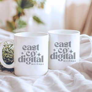 Puede incluir: Dos tazas de cerámica blanca con texto negro que dice "east co digital mockups" sobre un fondo de tela blanca.