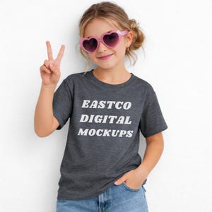 May include: A young person wearing a dark gray t-shirt with the text "EASTCO DIGITAL MOCKUPS" in white. The person is also wearing pink heart-shaped sunglasses and blue jeans. The person is holding up two fingers in a peace sign.