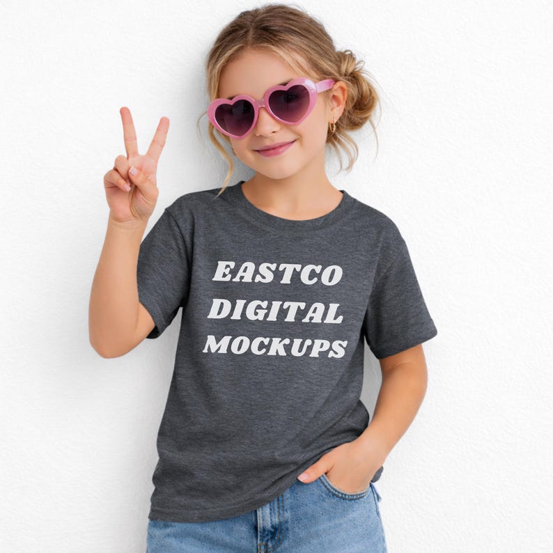 May include: A young person wearing a dark gray t-shirt with the text "EASTCO DIGITAL MOCKUPS" in white. The person is also wearing pink heart-shaped sunglasses and blue jeans. The person is holding up two fingers in a peace sign.