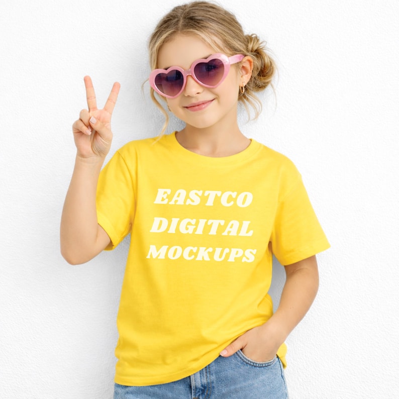 May include: A young person wearing a yellow t-shirt with the text "EASTCO DIGITAL MOCKUPS" in white. They are also wearing pink heart-shaped sunglasses and blue jeans. The person is holding up two fingers in a peace sign.