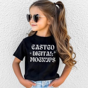 Könnte beinhalten: Ein schwarzes T-Shirt mit dem Text "EASTCO DIGITAL MOCKUPS" in einer Retro-Schriftart. Das Shirt wird von einer jungen Person mit langen braunen Haaren und herzförmigen Sonnenbrillen getragen.