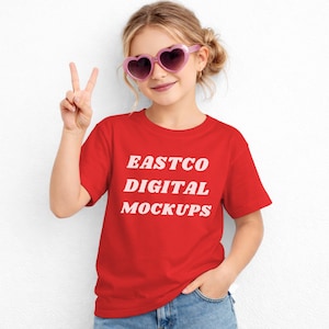 May include: A young person wearing a red t-shirt with the text "EASTCO DIGITAL MOCKUPS" in white. They are also wearing pink heart-shaped sunglasses and blue jeans. The person is holding up two fingers in a peace sign.