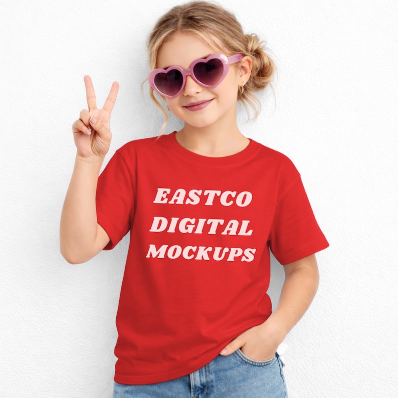 May include: A young person wearing a red t-shirt with the text "EASTCO DIGITAL MOCKUPS" in white. They are also wearing pink heart-shaped sunglasses and blue jeans. The person is holding up two fingers in a peace sign.